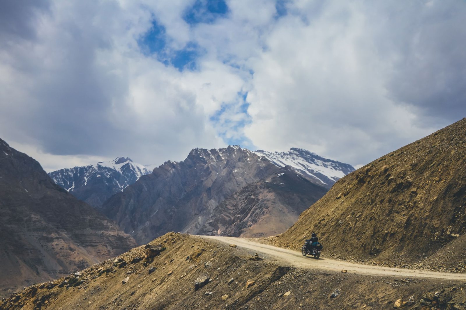 Scenic view of a motorcycle journey on Komic road amidst snow-capped mountains under a cloudy sky.