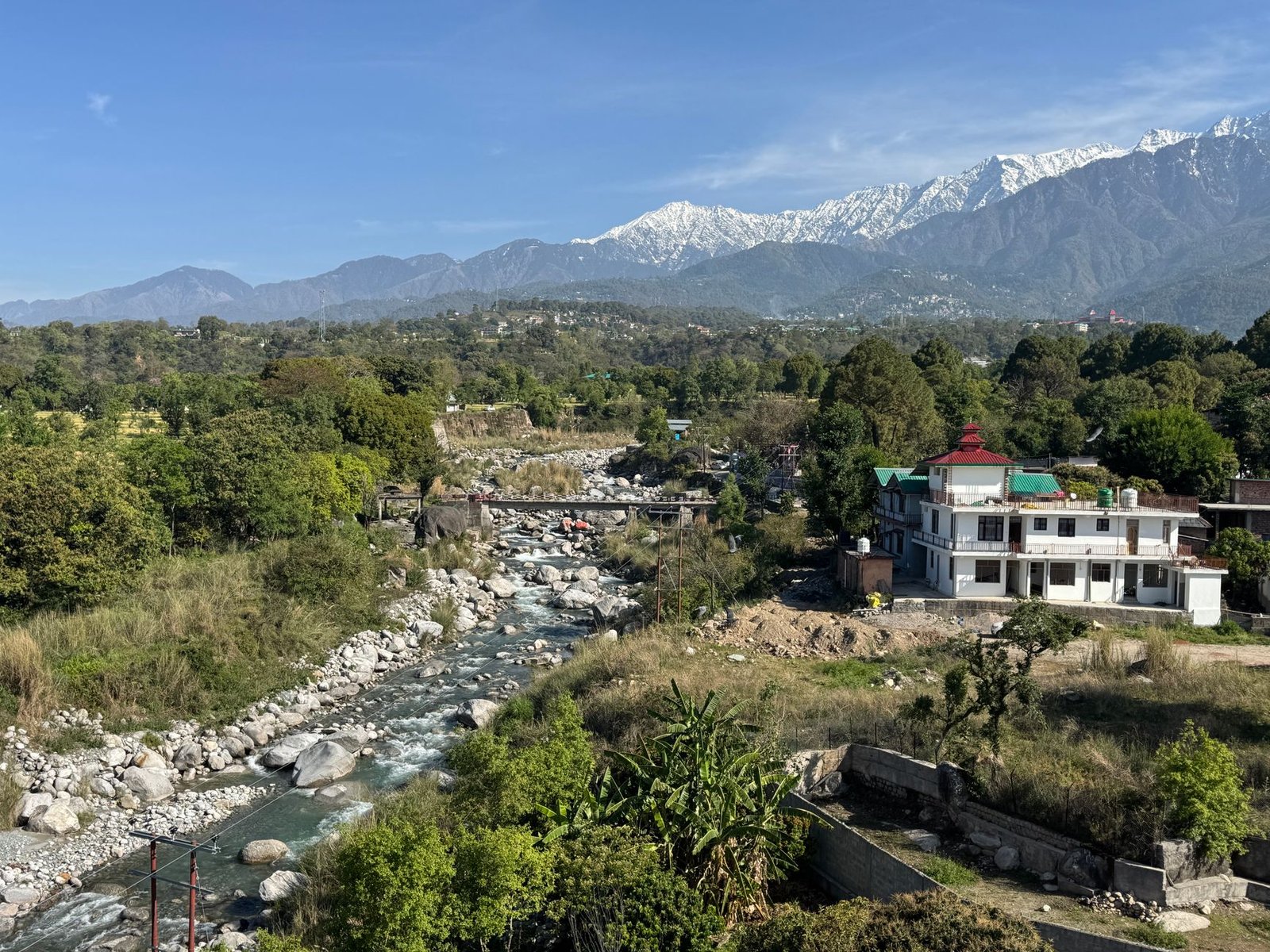 Picturesque landscape of Dharamshala, India showcasing a river, lush greenery, and snowy mountains.