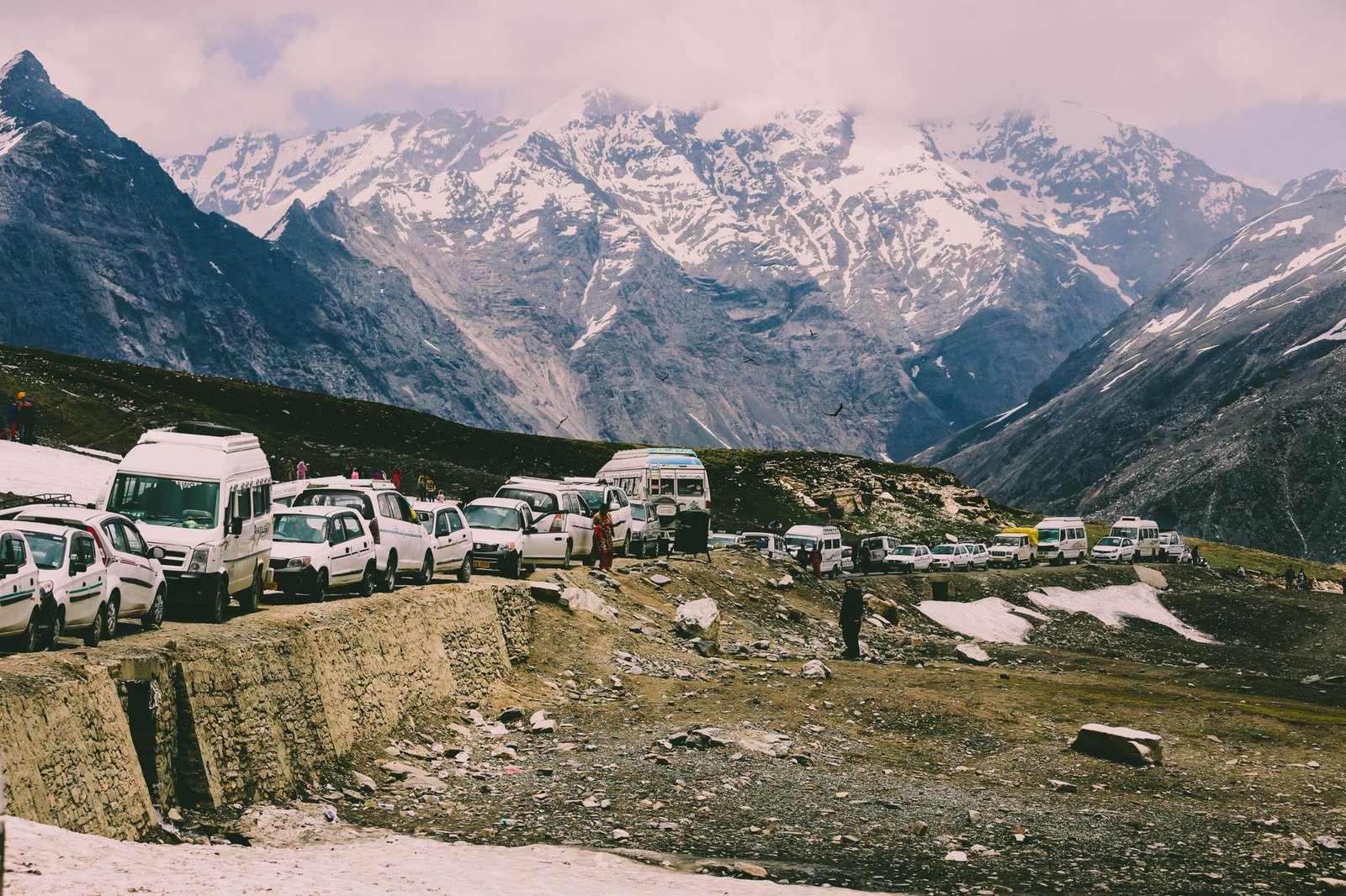 Vehicles lined up on a snowy mountain road, surrounded by majestic peaks and clear sky.