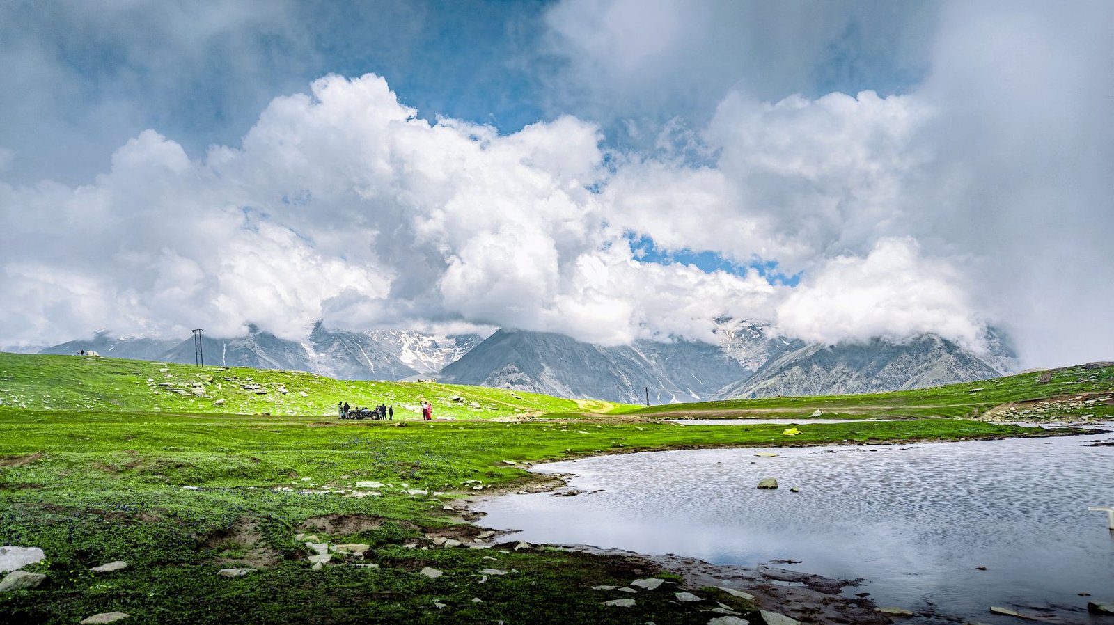 A breathtaking view of the Himalayas with lush green fields, a pristine lake, and dramatic clouds overhead.