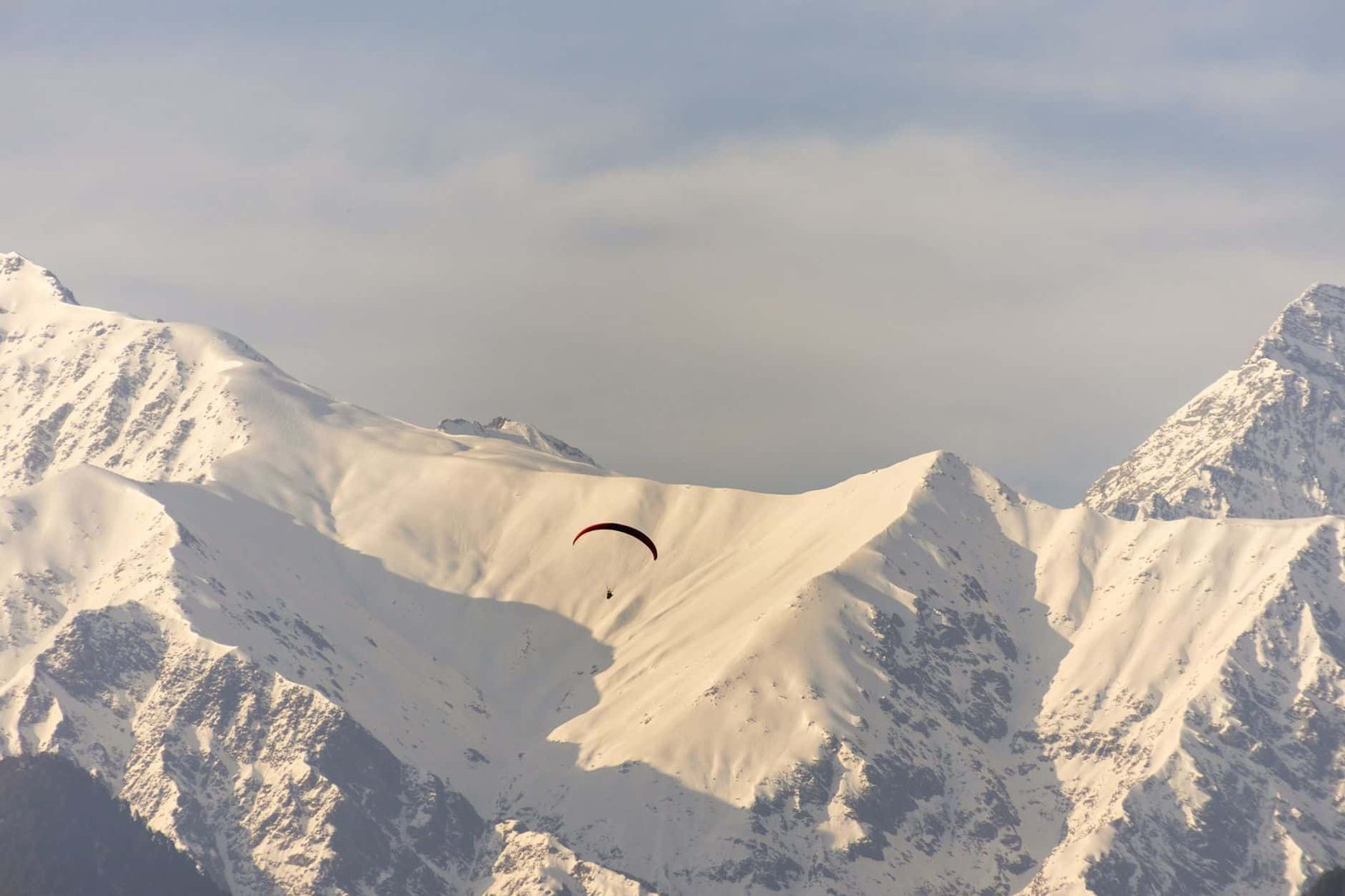 A paraglider soars over the majestic, snow-capped Dhauladhar mountains in Baijnath, Himachal Pradesh.